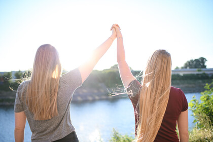Zwei junge Frauen sind von hinten zu sehen. Sie schauen auf einen Fluss und reichen sich in der Luft die Hand.