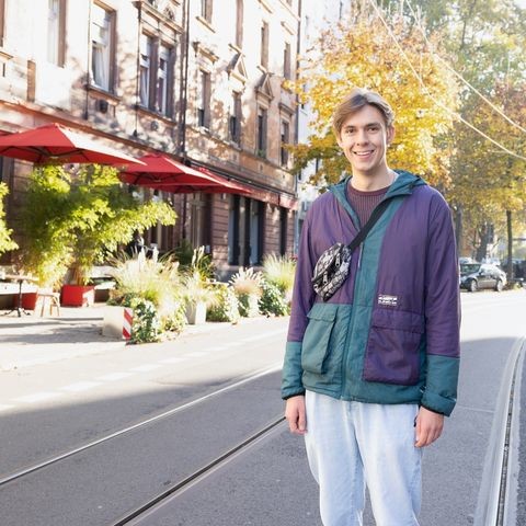 Porträt von VWL-Student Tim Bartelsen. Bartelsen trägt eine grün-violette Outdoorjacke, eine Gürteltasche über der Schulter und weite Jeans. Er steht auf den Schienen der Seckenheimer Landstraße in der Schwetzinger Vorstadt und lacht offenen in die Kamera. Link: Instagram-Post Stadtteile Mannheims