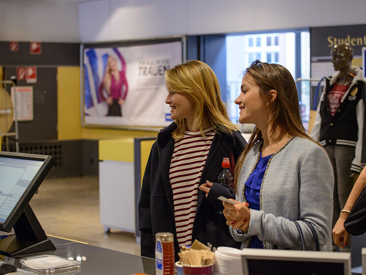 Zwei Frauen stehen vor dem Uni-Shop. Die Frau links hat lange blonde Haare und trägt ein gestreiftes Oberteil mit einer blauen Jacke darüber. Die Frau rechts daneben hat lange braune Haare und trägt ein blaues Oberteil mit einer grauen Jacke darüber.