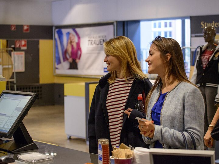 Zwei Frauen stehen vor dem Uni-Shop. Die Frau links hat lange blonde Haare und trägt ein gestreiftes Oberteil mit einer blauen Jacke darüber. Die Frau rechts daneben hat lange braune Haare und trägt ein blaues Oberteil mit einer grauen Jacke darüber.