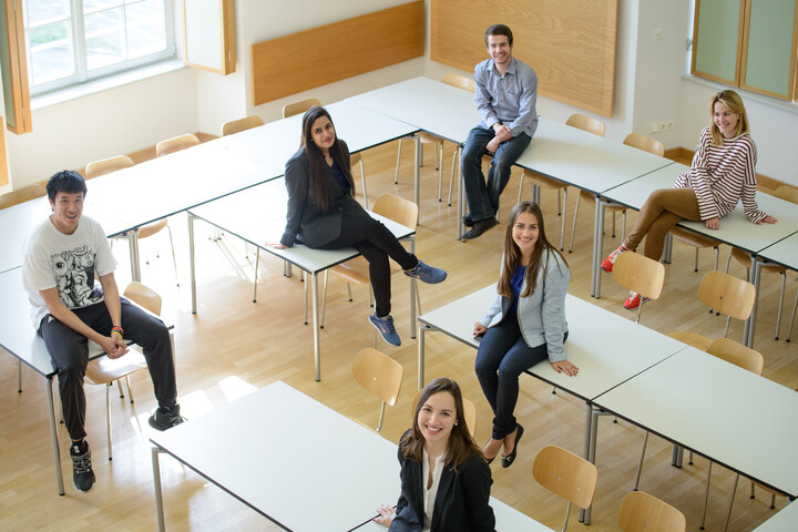 Von oben aufgenommene Gruppe von sechs Studierenden, die auf Tischen in einem hellen Seminarraum sitzen.