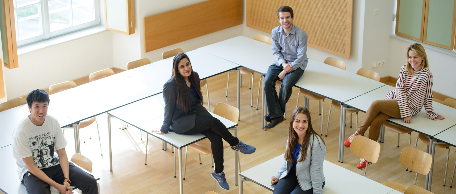 Von oben aufgenommene Gruppe von sechs Studierenden, die auf Tischen in einem hellen Seminarraum sitzen.