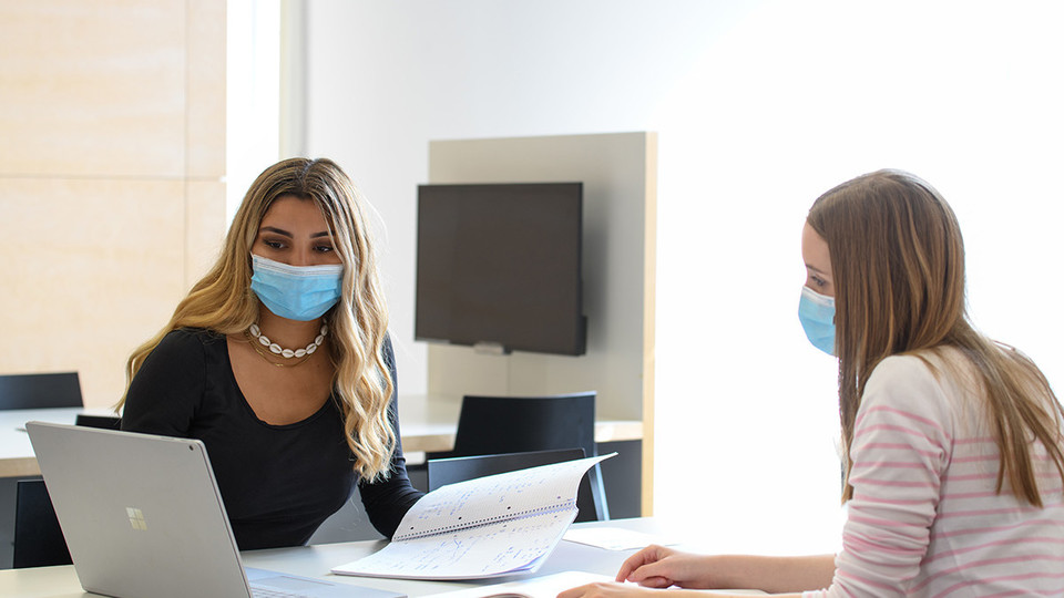 Zwei Frauen sitzen sich an einem Schreibtisch gegenüber. Beide tragen eine medizinische Maske und schauen auf einen Laptop.