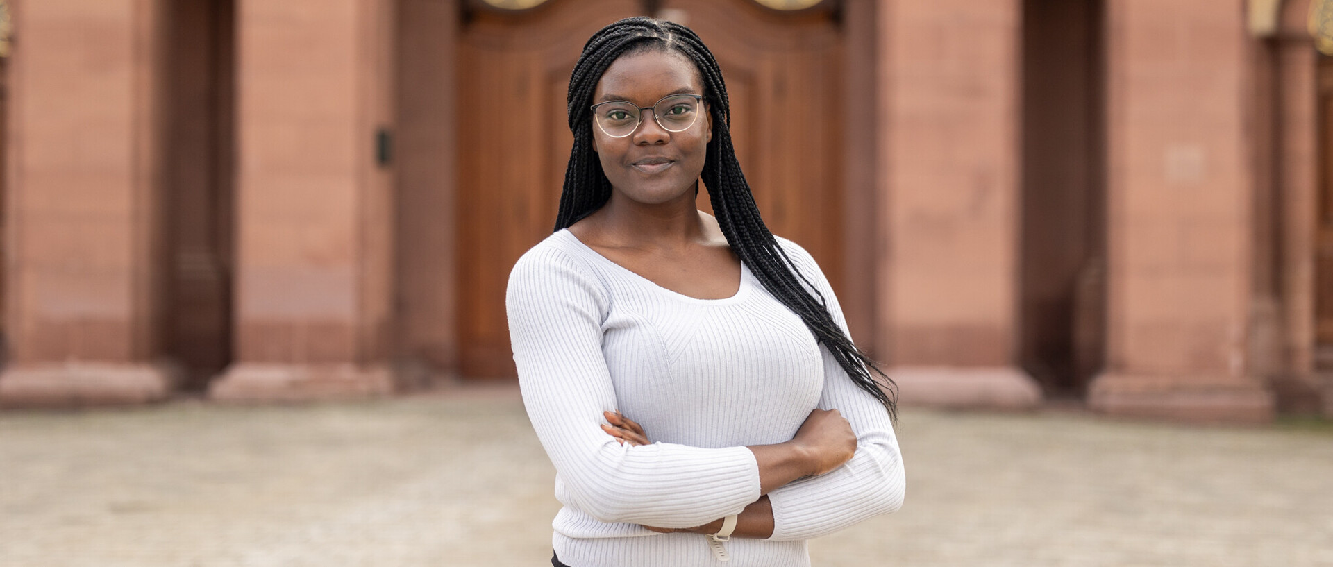 Halkörperportrait von Esther mit locker verschränkten Armen vor dem Schloss. Sie trägt schwarze lange Braids, eine Brille und einen weißen Pullover.