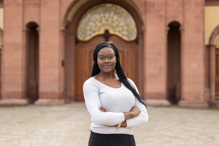 Halkörperportrait von Esther mit locker verschränkten Armen vor dem Schloss. Sie trägt schwarze lange Braids, eine Brille und einen weißen Pullover.