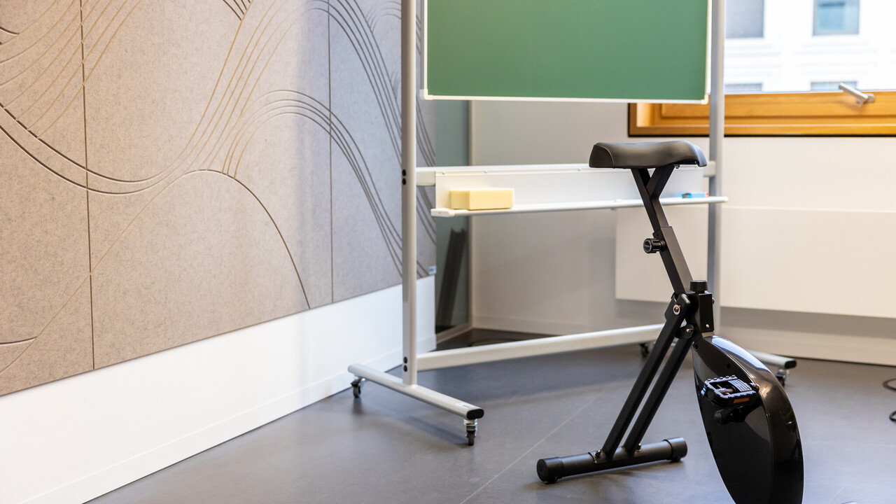 Das Bild zeigt ein Sitzfahrrad vor einer Kreidetafel. Links ist eine schallschluckende hellbraune Wand zu sehen. Im Hintergrund der Teil eines Fensters.