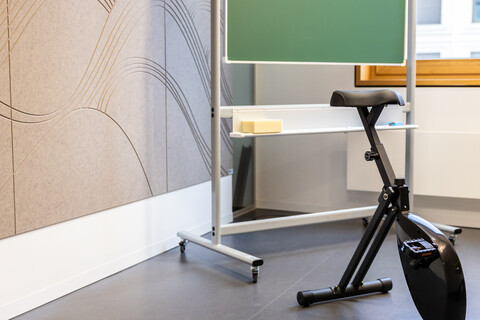 Das Bild zeigt ein Sitzfahrrad vor einer Kreidetafel. Links ist eine schallschluckende hellbraune Wand zu sehen. Im Hintergrund der Teil eines Fensters.