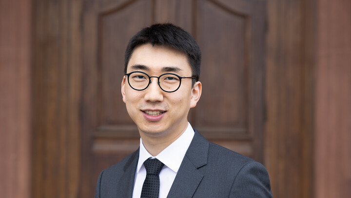 Eunseong Park. He is wearing a black suit with a black tie and is standing in front of a brown door.