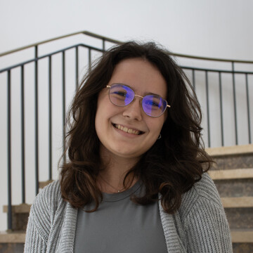 Vanessa trägt eine Brille und hat schulterlange, dunkel gelockte Haare. Sie trägt eine hellgraue Strickjacke und lächelt freundlich in die Kamera.