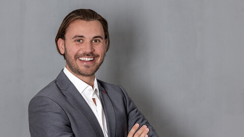 Cedric Stricker. He is wearing a grey suit and is standing in front of a grey wall.