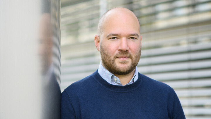 Richard Traunmüller blickt ernst in die Kamera. Er trägt einen blauen Pullover und lehnt sich an eine graue Wand. Im Hintergrund sind Lamellen eines Rolladens zu sehen.