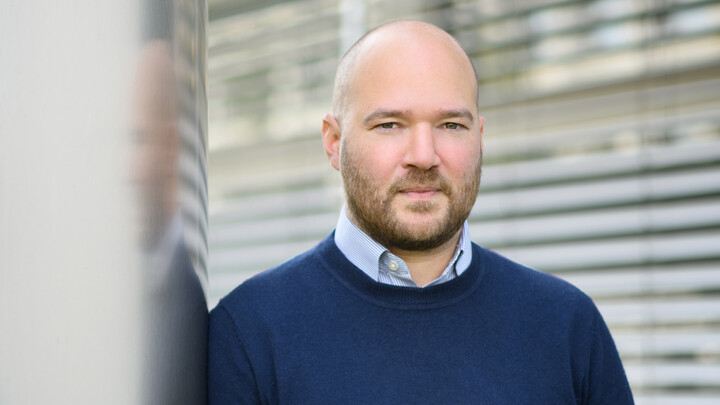 Richard Traunmüller blickt ernst in die Kamera. Er trägt einen blauen Pullover und lehnt sich an eine graue Wand. Im Hintergrund sind Lamellen eines Rolladens zu sehen.