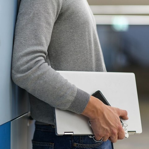 Ein Mann lehnt mit dem Rücken an einer hellblauen Wand. Er trägt einen grauen Pulli und einen Laptop unter dem Arm. Link: Instagram Post ENGAGE.EU