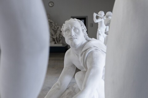 Eine Skulptur eines niedergeknieten Mannes blickt nach oben auf eine weitere Skulptur, deren Glieder im Vordergrund zu sehen sind.