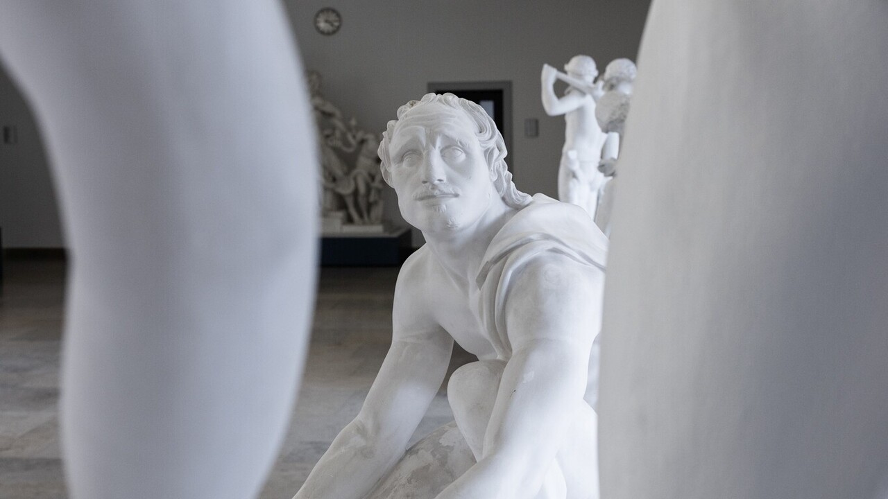 Eine Skulptur eines niedergeknieten Mannes blickt nach oben auf eine weitere Skulptur, deren Glieder im Vordergrund zu sehen sind.