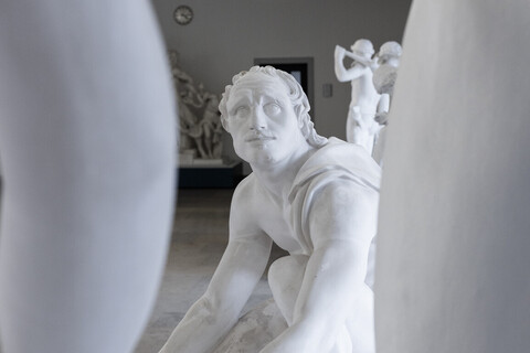 Eine Skulptur eines niedergeknieten Mannes blickt nach oben auf eine weitere Skulptur, deren Glieder im Vordergrund zu sehen sind.