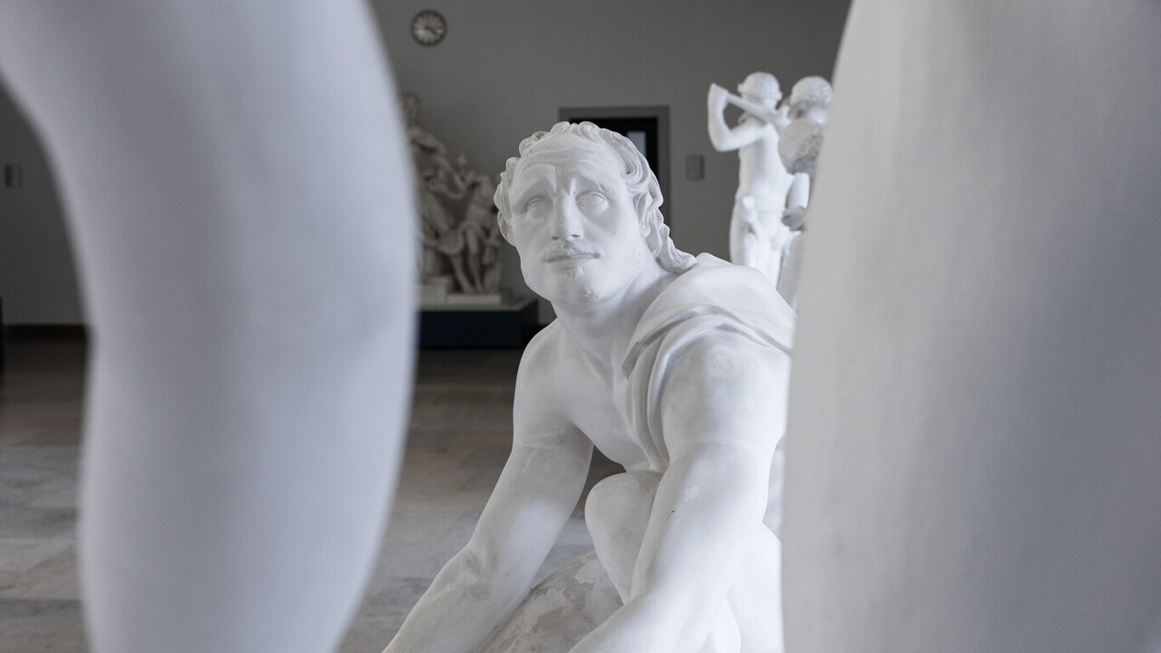 Eine Skulptur eines niedergeknieten Mannes blickt nach oben auf eine weitere Skulptur, deren Glieder im Vordergrund zu sehen sind.