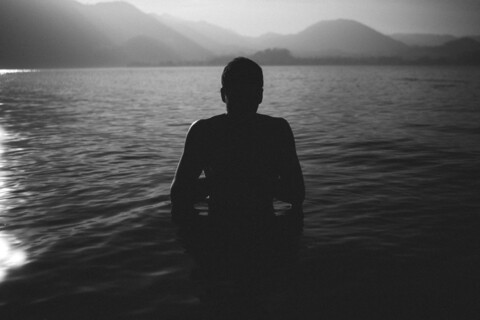 Schwarz-weiß Foto: Eine Person steht bis zur Hüfte in einem See umgeben von Bergen, das Gesicht ist durch Schatten verdeckt.