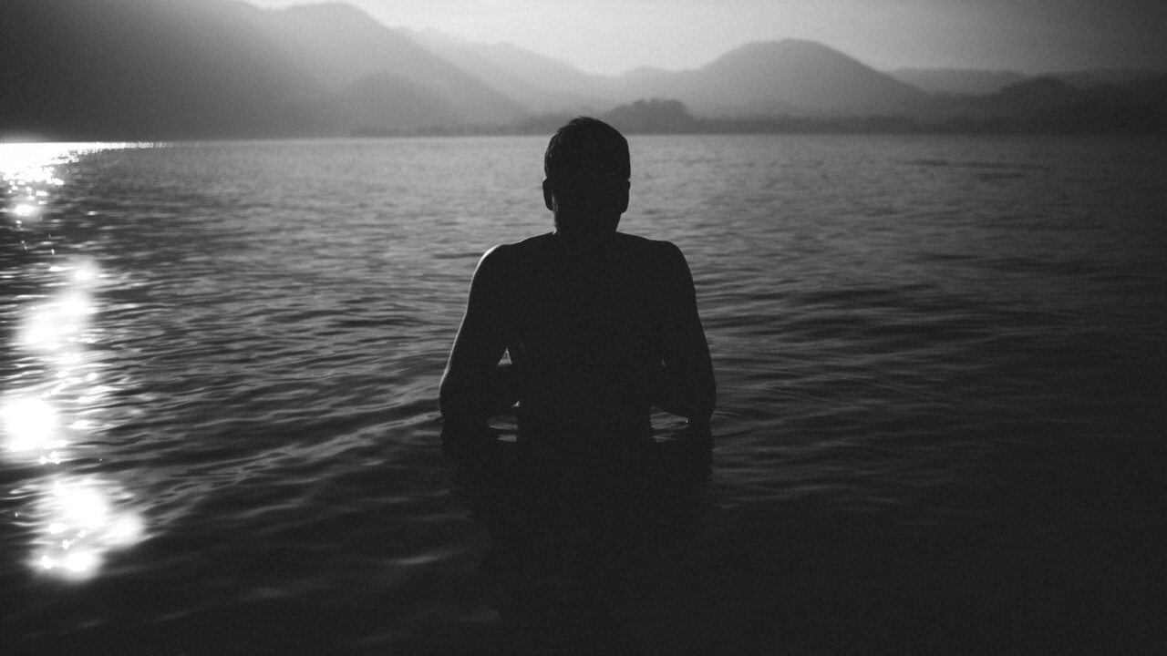 Schwarz-weiß Foto: Eine Person steht bis zur Hüfte in einem See umgeben von Bergen, das Gesicht ist durch Schatten verdeckt.