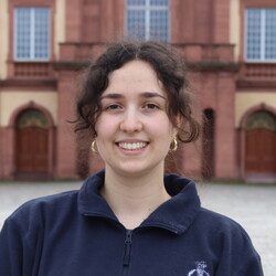 Lilli Hirt. Die Person steht vor dem Schloss, trägt einen dunkelblauen Pullover und lächelt in die Kamera.