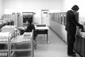 schwarz-weiß Aufnahme des damaligen Maschinensaals. Eine Person sitzt an einem Tisch und die andere steht vor einer Maschine. 