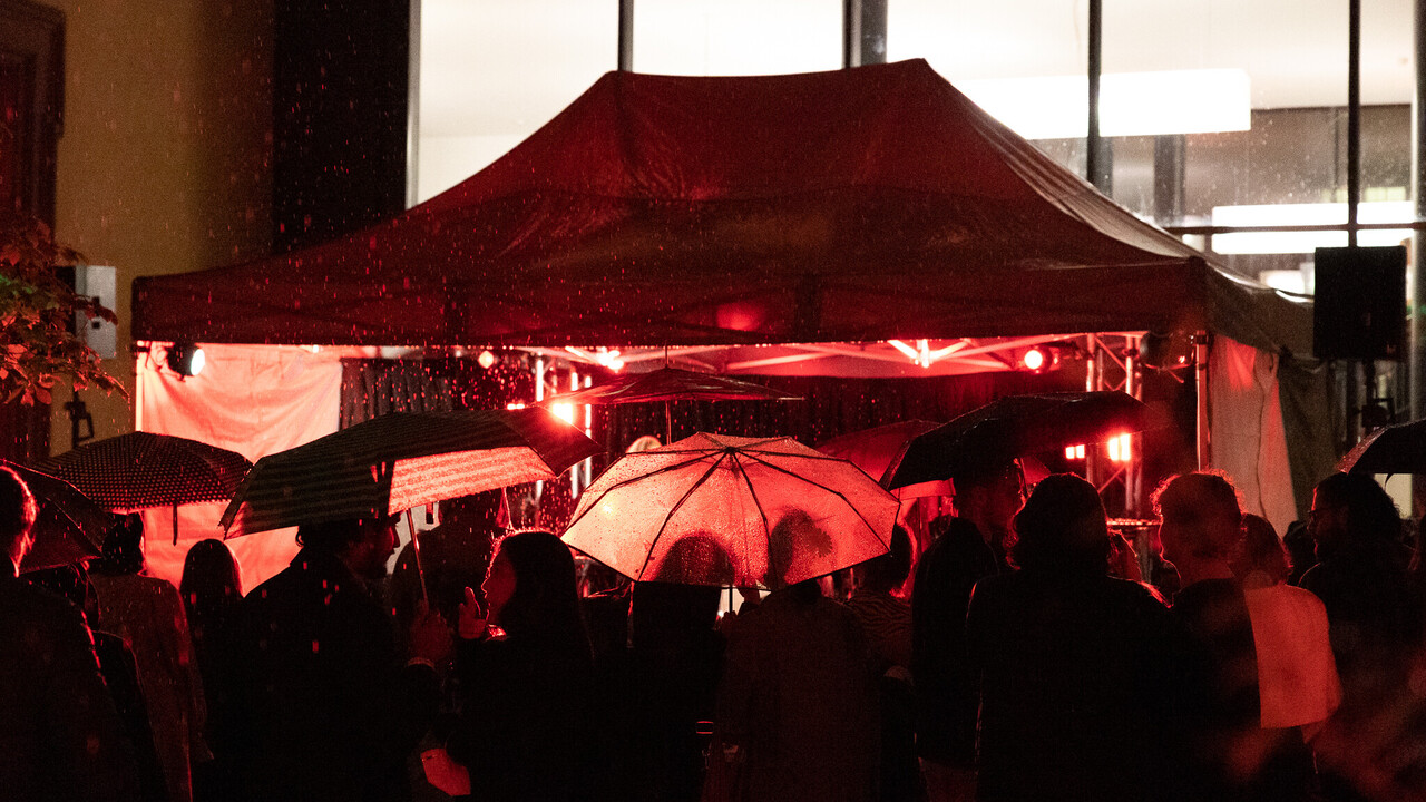 Eine Menschenmenge schützt sich unter Schirmen und einem Zelt vor einem Regenschauer.