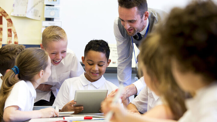 Schüler*innen sitzen an einem Tisch und ein Lehrer erklärt ihnen etwas.