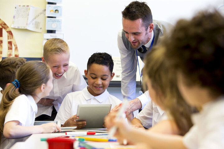 Schüler*innen sitzen an einem Tisch und ein Lehrer erklärt ihnen etwas.