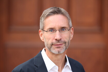 Portrait von Jürgen Seifried. Er trägt ein weißes Hemd sowie einen schwarzen Blazer und steht vor einer Holztüre.