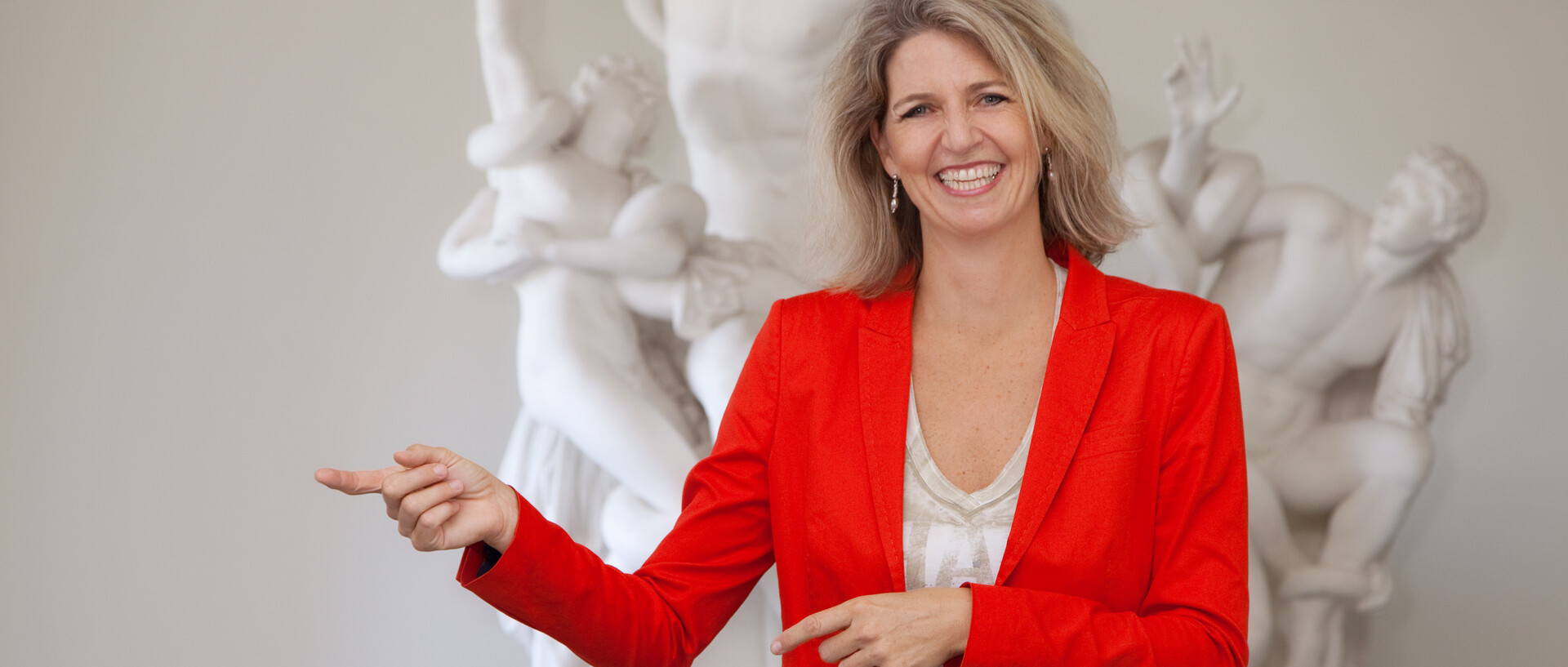 Eine Frau in rotem Blazer zeigt nach rechts. Im Hintergrund ist eine Statue.