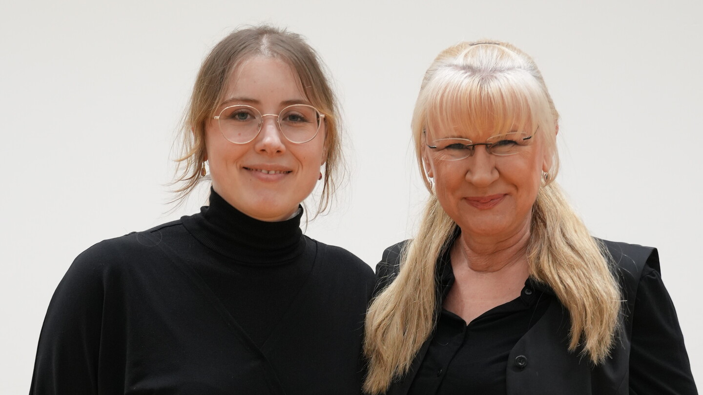 Zwei schwarz gekleidete Menschen stehen nebeneinander vor einer weißen Wand und lächeln in die Kamera.