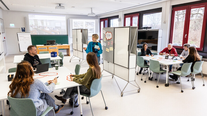 Mehrere Personen sitzen an zwei Toschen im EduSpace. Eine Person steht an einer Tafel.