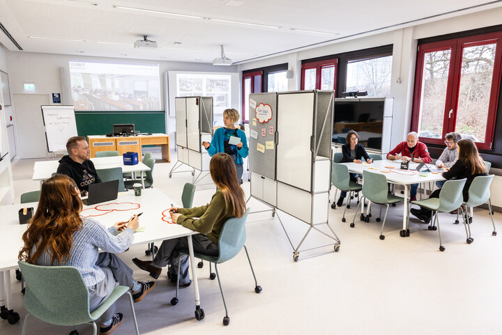 Mehrere Personen sitzen an zwei Toschen im EduSpace. Eine Person steht an einer Tafel.