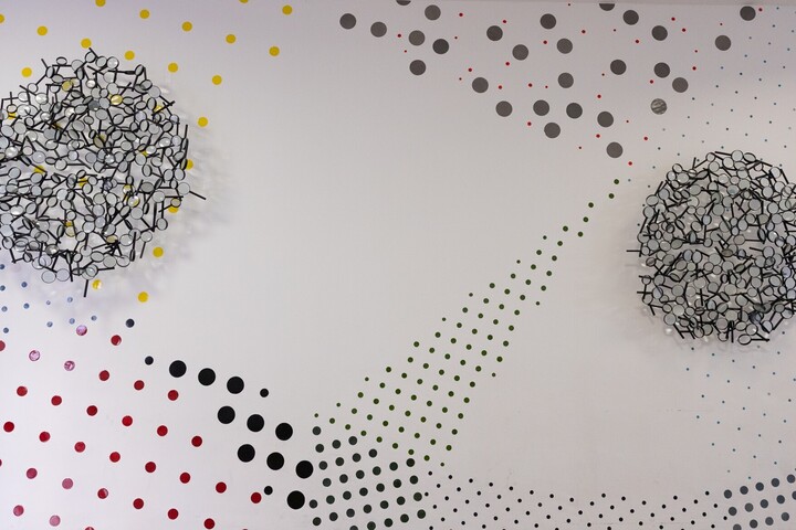 Eine Wand in einer Galerie mit zwei abstrakten, kugelförmigen Skulpturen aus schwarzem und weißem Draht, die vor einem Hintergrund aus farbigen Punkten in Gelb, Rot, Schwarz und Grau hängen. Die Punkte variieren in Größe und Dichte und bilden fließende Muster auf der weißen Wandfläche.