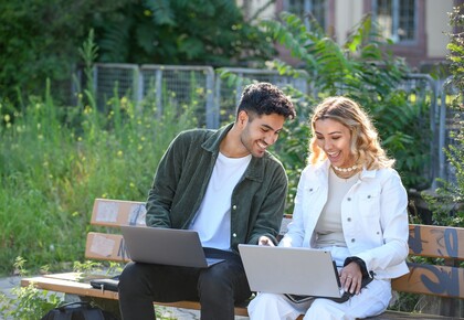 Zwei Studierende sitzen mit ihren Laptops auf einer Bank im Grünen. Sie lächeln.