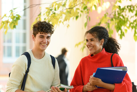 Ein Student und eine Studentin stehen im Hof des Schlosses. Er trägt einen Rucksack, sie hält einen Ordner im Arm und hat eine Tasche über der Schulter.