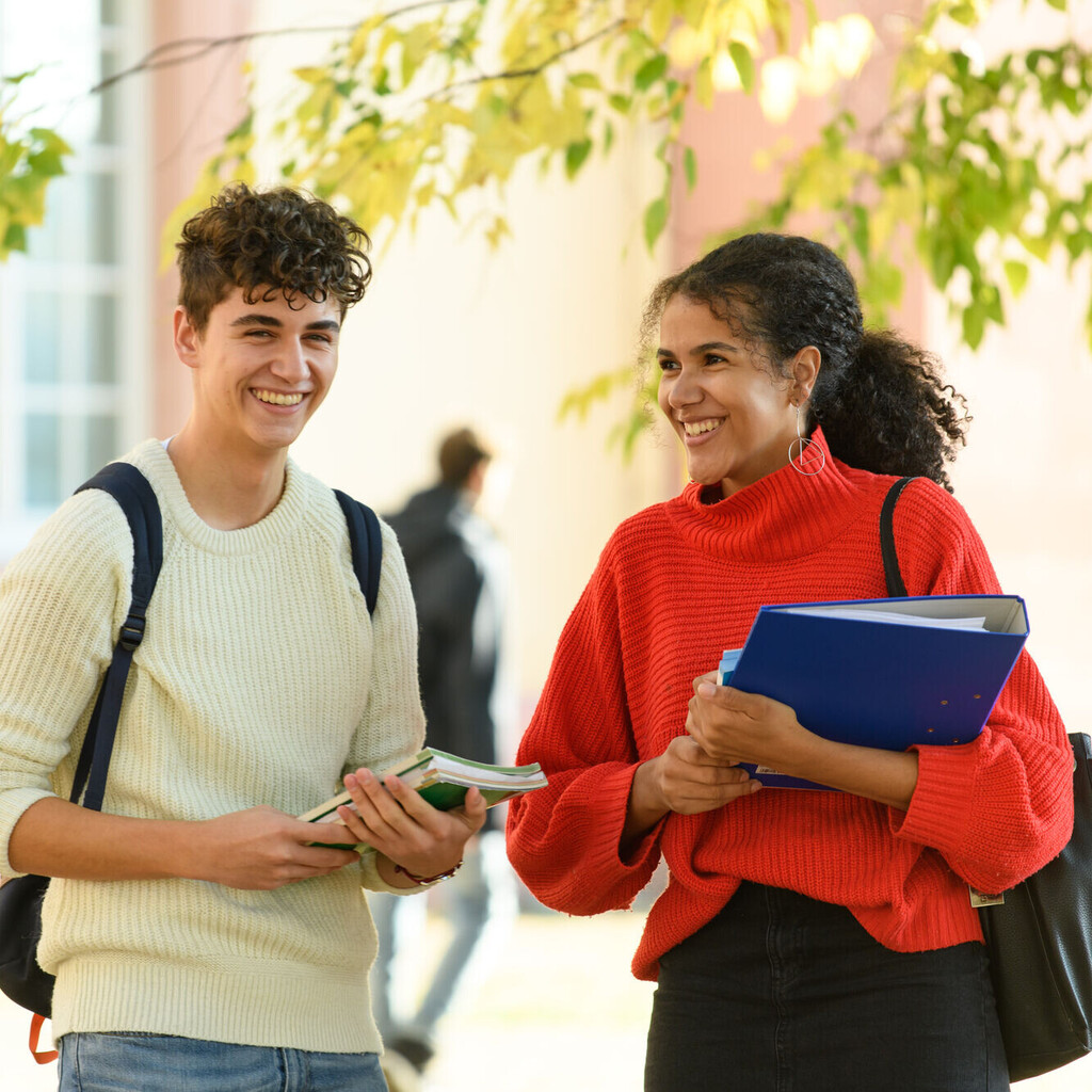 Ein Student und eine Studentin stehen im Hof des Schlosses. Er trägt einen Rucksack, sie hält einen Ordner im Arm und hat eine Tasche über der Schulter.