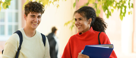Ein Student und eine Studentin stehen im Hof des Schlosses. Er trägt einen Rucksack, sie hält einen Ordner im Arm und hat eine Tasche über der Schulter.