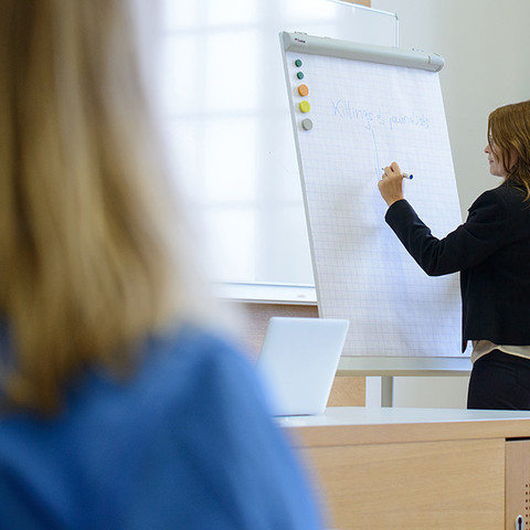 Eine Dozentin schreibt mit blauem Stift auf ein Flipchart "Killings of journalists", während im Vordergrund eine Studentin in Rückenansicht sitzt.