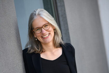Portrait von Jutta Mata. Sie trägt einen schwarzen Pullover sowie einen schwarzen Cardigan und steht lächelnd an einer Wand.