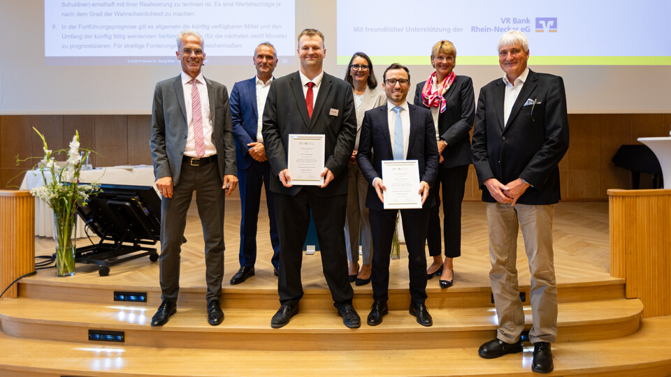 Gruppenbild mit den Preisträgern Dr. Johannes Heck und Dr. Ansgar Kalle mit den Mitglieder der Auswahlkommission Prof. Dr. Georg Bitter, RA Tobias Hoefer, Vors. RiBGH a.D. Prof. Dr. Godehard Kayser, RAin Annette Kollmar, RAin Dr. Alexandra Schluck-Amend. Alle Personen sind mit Anzügen gekleidet und stehn auf einer Bühne. Die beiden Preisträger zeigen ihre Urkunden.
