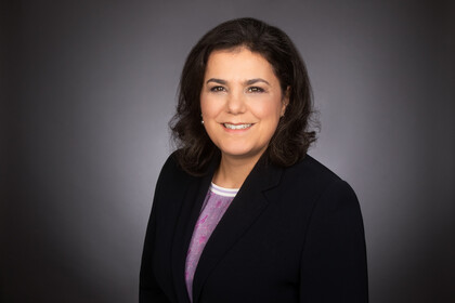 Portrait von Carmela Aprea. Sie trägt einm lila Shirt und einen schwarzen Blazer und steht lächelnd vor einer grauen Wand.