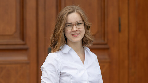 Nina Osenbrügge. She is wearing a white shirt and is standing in front of a brown door.