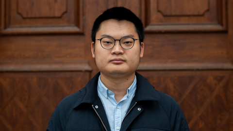 Quinkun Yao. He is wearing a black jacket over a blue shirt and is standing in front of brown door.