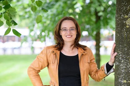 Portrait von Michele Tertilt. Sie trägt einen schwarzen Pollover sowie eine orange farbige Jacke und steht auf einer Grünfäche.