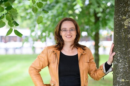 Portrait von Michele Tertilt. Sie trägt einen schwarzen Pollover sowie eine orange farbige Jacke und steht auf einer Grünfäche.