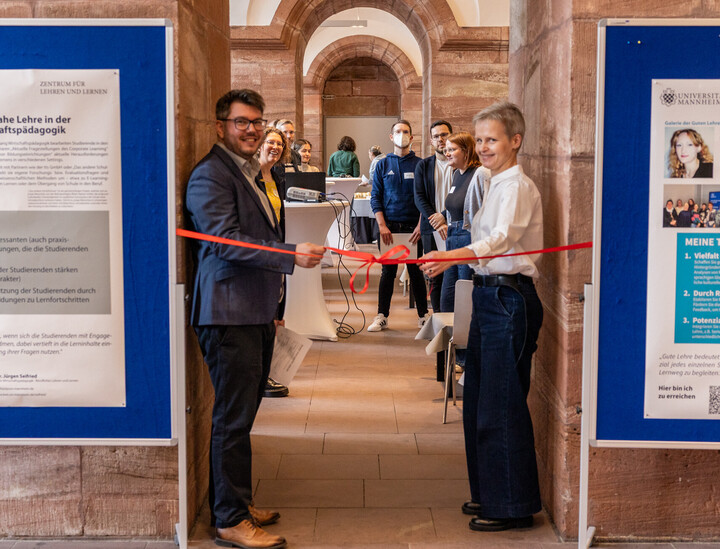 Feierliche Eröffnung in den Katakomben der Universität: Zwei Personen durchtrennen ein rotes Band zur Eröffnung der Galerie der guten Lehre. Im Hintergrund warten mehrere Personen darauf, die Galerie betreten zu können.