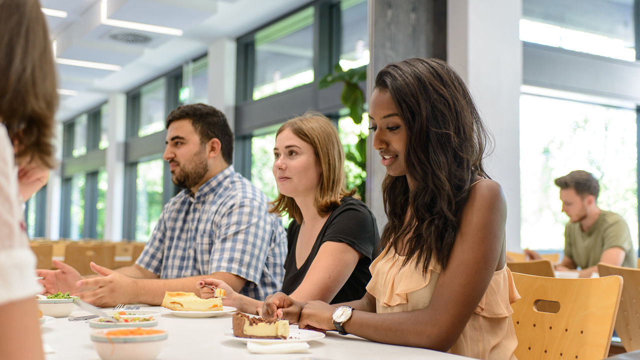 Studierende sitzen in der Mensa an einem Tisch und essen gemeinsam zu Mittag. Zwei Studentinnen essen Kuchen.