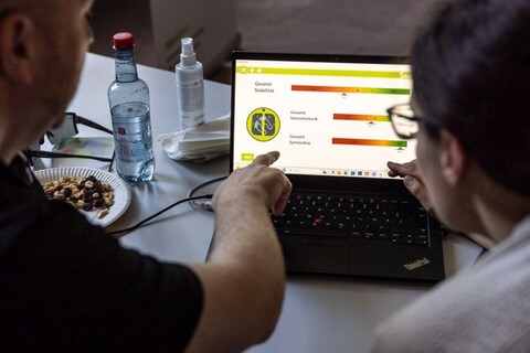Auswertung eines Balance-Experiments. Mann und Frau schauen auf Bildschirm eines Laptops. Auf Bildschirm zwei Skalas zur Auswertung von "Gesamtsensomotorik" und "Gesamtsymmetrie".