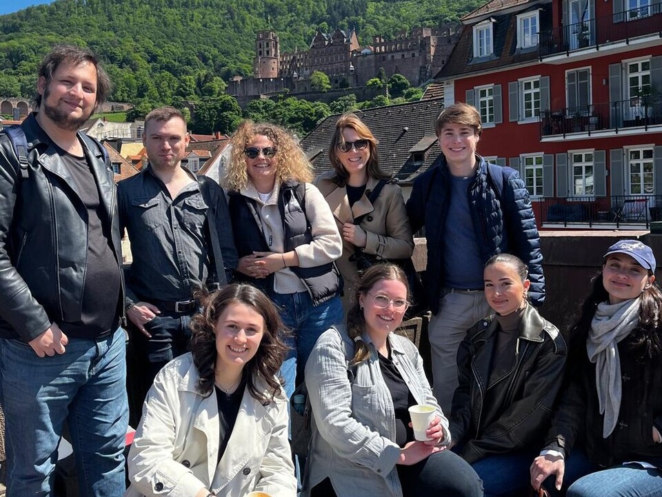 Eine Gruppe von neun Personen auf der Alten Brücke in Heidelberg, im Hintergrund rechts ein rotes Haus und die Ruine des Heidelberger Schlosses.